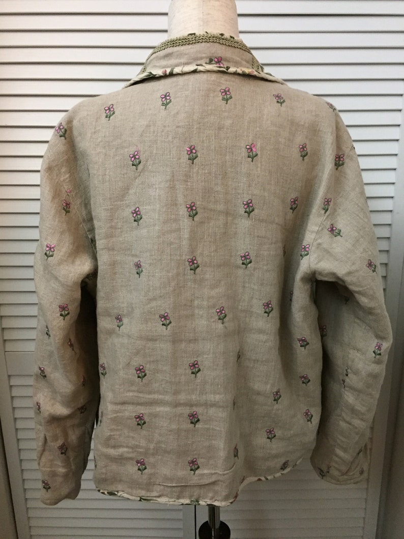 May include: A beige linen shirt with long sleeves and a floral pattern. The flowers are embroidered in pink and green.