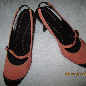 May include: A pair of coral pink suede heeled shoes with black trim and a black strap across the foot.