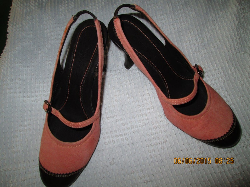May include: A pair of coral pink suede heeled shoes with black trim and a black strap across the foot.