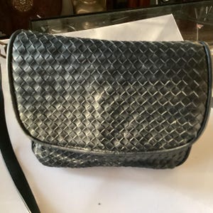 Könnte beinhalten: Eine schwarze Leder-Umhängetasche mit gewebtem Muster. Die Tasche hat einen langen Riemen und einen Klappenverschluss.