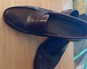 May include: A pair of dark brown leather penny loafers featuring a classic moc-toe construction with a stitched apron and a traditional strap across the vamp. The interior footbed is dark brown with perforated detailing and the Bass & Co. logo.