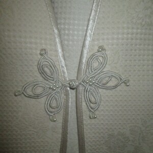 May include: A white garment with a decorative knot closure featuring a series of loops and white beads.