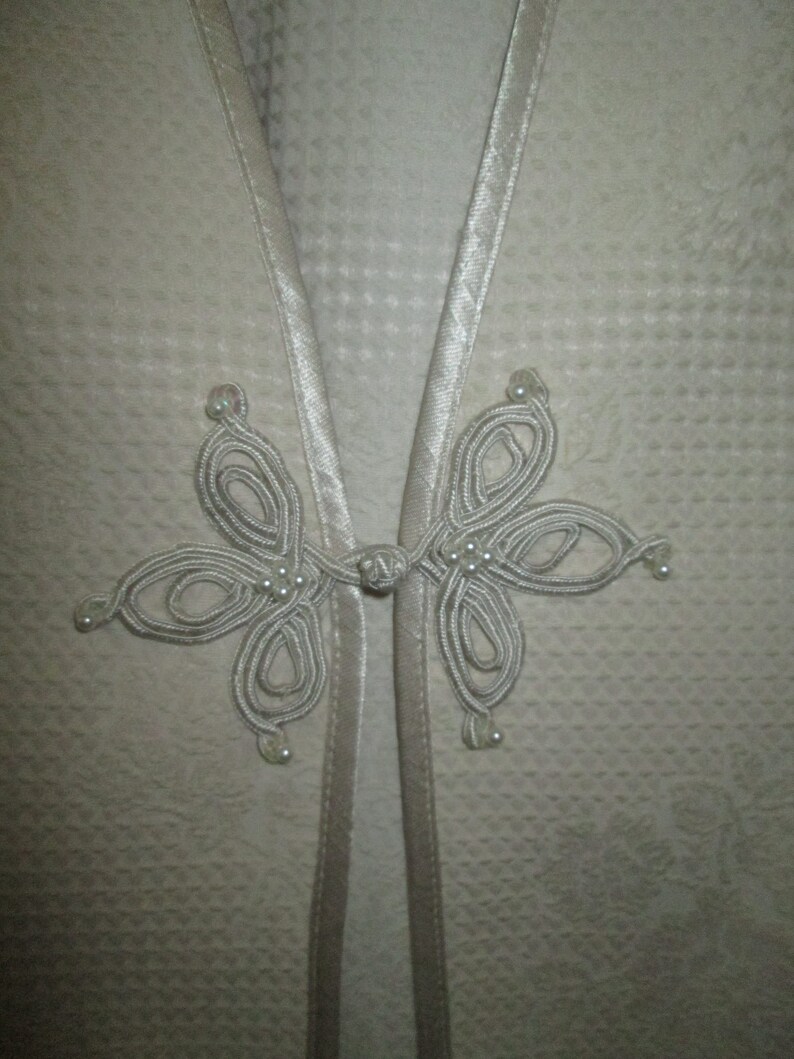 May include: A white garment with a decorative knot closure featuring a series of loops and white beads.