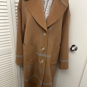 May include: A long, tan coat with a classic collar and three decorative buttons. The coat features two front pockets and gray stripes on the sleeves and lower hem. A patterned scarf is visible inside the coat.