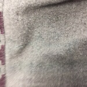 May include: Close-up of a light gray fabric with a subtle texture. The fabric has a border with a geometric pattern in shades of red, green, and purple.