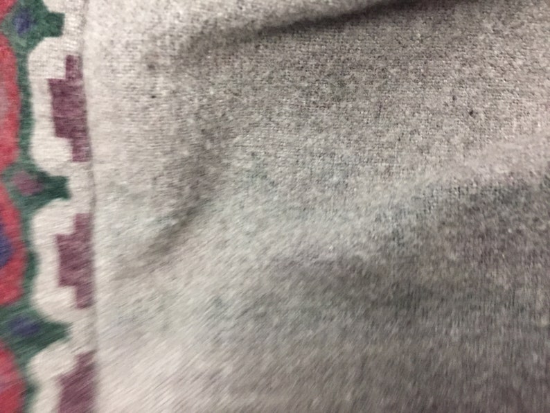 May include: Close-up of a light gray fabric with a subtle texture. The fabric has a border with a geometric pattern in shades of red, green, and purple.