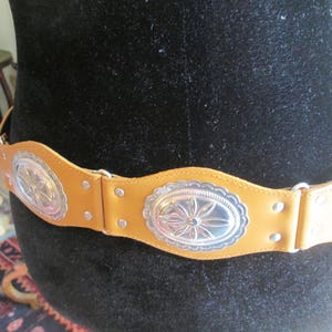 May include: A brown leather belt with silver concho accents. The belt has three oval-shaped conchos with a floral design.