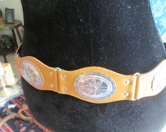 May include: A brown leather belt with silver concho accents. The belt has three oval-shaped conchos with a floral design.