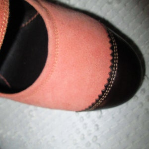May include: A close-up of a pink suede shoe with a black leather toe cap and a decorative stitch detail. The shoe has a strap across the top.