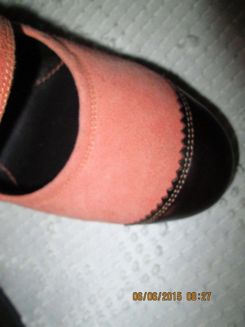 May include: A close-up of a pink suede shoe with a black leather toe cap and a decorative stitch detail. The shoe has a strap across the top.