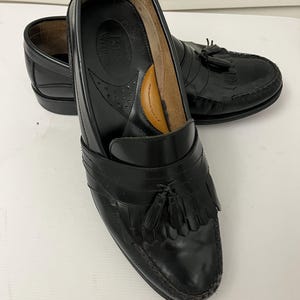 May include: Black leather loafers with tassel detailing. The shoes have a classic design with a rounded toe and a low heel. The interior features a brown leather insole. The shoes are in good condition and appear to be well-maintained.