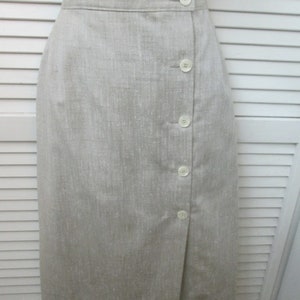 May include: A knee-length skirt in a light beige, textured fabric. The skirt features a high waistband and a vertical front closure with five round, off-white buttons arranged in a single column along the right side of the front panel.