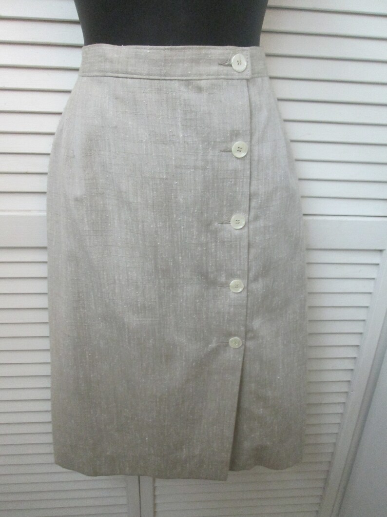 May include: A knee-length skirt in a light beige, textured fabric. The skirt features a high waistband and a vertical front closure with five round, off-white buttons arranged in a single column along the right side of the front panel.