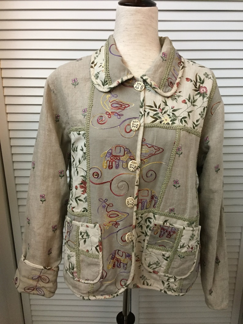 May include: A beige linen jacket with a patchwork design featuring embroidered birds, elephants, and floral patterns. The jacket has a collared neckline, long sleeves, and button closures.