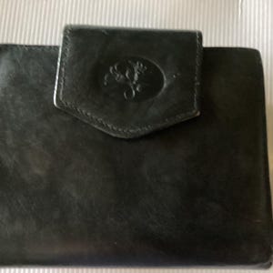 May include: A black leather wallet with a flap closure. The flap features an embossed design within an oval shape. The wallet is rectangular and appears to be made of a smooth, durable material. The overall design is simple and classic.