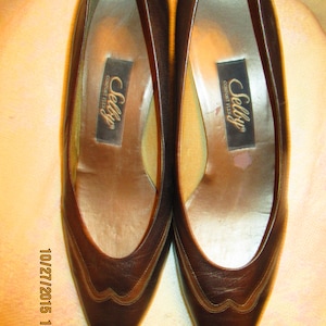 May include: A pair of brown leather women's shoes with a pointed toe and a small heel. The shoes have a decorative stitching detail on the toe and are labeled "Selby Comfort Flex".