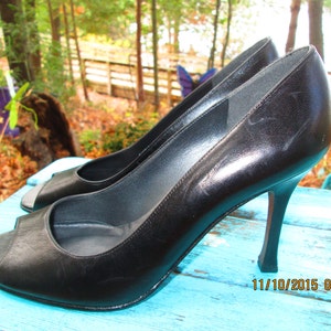 May include: A pair of black leather peep-toe heels with a stiletto heel. The shoes are on a blue wooden surface.
