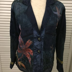 Puede incluir: Una chaqueta azul de manga larga con un diseño de patchwork que presenta motivos florales y de hojas en tonos naranja, verde y marrón. La chaqueta tiene cuello en V, cierre de botón y dos bolsillos.