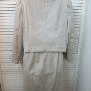 May include: A beige two-piece set consisting of a cropped long-sleeve jacket with a collarless neckline and a matching knee-length skirt. The garment features a subtle vertical textured weave throughout, providing a consistent neutral tone.