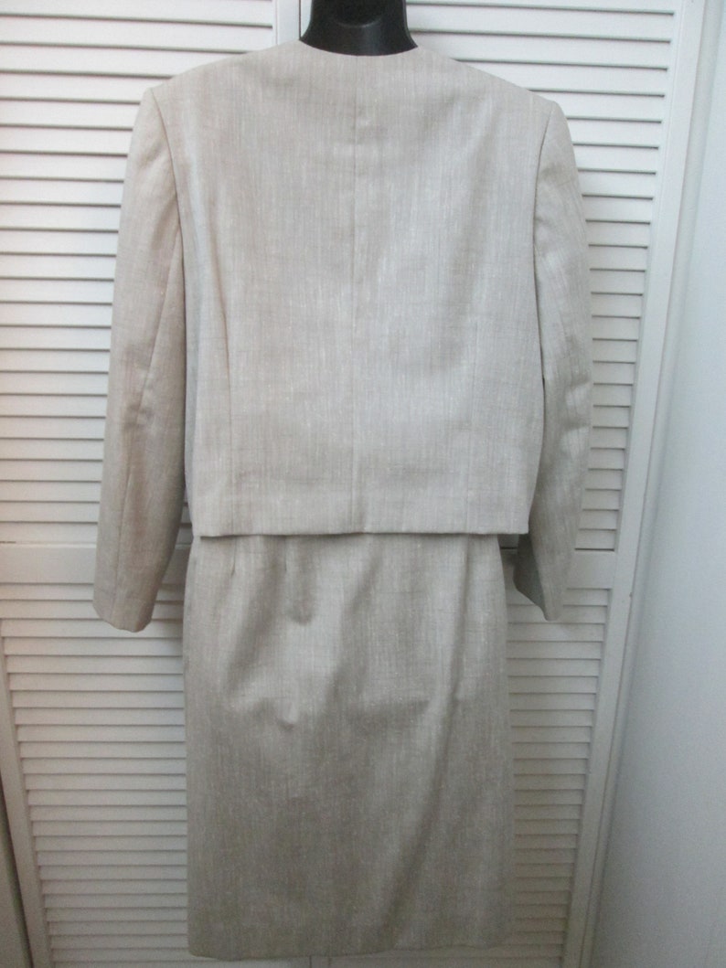 May include: A beige two-piece set consisting of a cropped long-sleeve jacket with a collarless neckline and a matching knee-length skirt. The garment features a subtle vertical textured weave throughout, providing a consistent neutral tone.