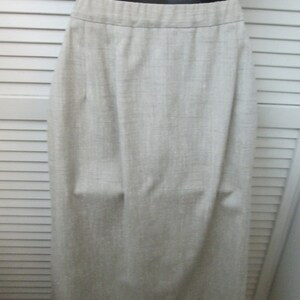 May include: A light beige, knee-length skirt with a visible woven texture. The garment features a simple, straight-cut silhouette with a clean, flat waistband and subtle vertical folds that drape naturally from the waist down to the hemline.