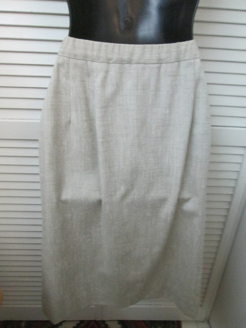 May include: A light beige, knee-length skirt with a visible woven texture. The garment features a simple, straight-cut silhouette with a clean, flat waistband and subtle vertical folds that drape naturally from the waist down to the hemline.