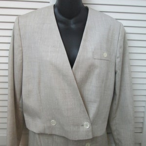 May include: A beige cropped blazer with long sleeves, a V-neckline, and a front button fastening. The jacket features a small chest pocket with a single button detail and a textured fabric finish. It is displayed on a black mannequin torso.