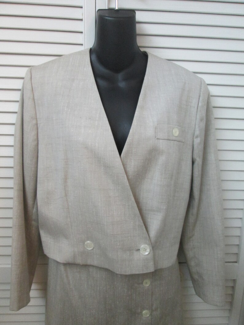 May include: A beige cropped blazer with long sleeves, a V-neckline, and a front button fastening. The jacket features a small chest pocket with a single button detail and a textured fabric finish. It is displayed on a black mannequin torso.