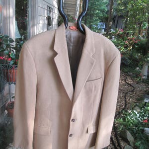 May include: A beige blazer with a three-button fastening and two pockets. The blazer is hanging on a black hanger.