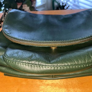May include: A dark forest green leather crossbody bag featuring a curved front flap with a brass-toned zipper closure and a matching brass-toned magnetic snap button. The bag has visible stitching along the edges and a leather shoulder strap.