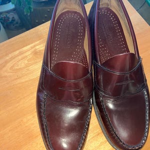 May include: A pair of burgundy leather penny loafers. The shoes have a classic design with a rounded toe, a strap across the vamp, and a stitched sole. The interior is a lighter shade of burgundy.