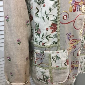 May include: A beige linen jacket with embroidered floral and geometric patterns. The jacket has a patch pocket on the front and a decorative trim around the edges.