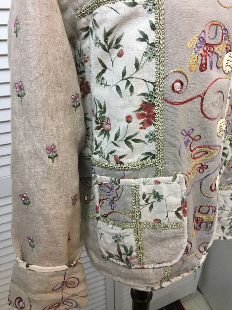 May include: A beige linen jacket with embroidered floral and geometric patterns. The jacket has a patch pocket on the front and a decorative trim around the edges.