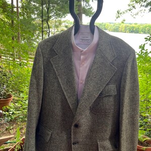 May include: A textured olive green and black blazer with a geometric diamond pattern, featuring notched lapels, two front buttons, and two lower patch pockets. Underneath is a light pink button-down shirt with a visible Calvin Klein label.