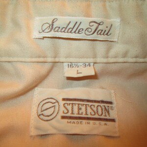 K&ouml;nnte beinhalten: Ein wei&szlig;es Hemd mit einem braunen "Saddle Tail"-Etikett und einem braunen "Stetson"-Etikett mit dem Text "Made in U.S.A." Das Gr&ouml;&szlig;enetikett lautet "16 1/2 - 34 L".