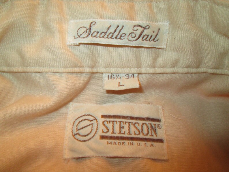 K&ouml;nnte beinhalten: Ein wei&szlig;es Hemd mit einem braunen "Saddle Tail"-Etikett und einem braunen "Stetson"-Etikett mit dem Text "Made in U.S.A." Das Gr&ouml;&szlig;enetikett lautet "16 1/2 - 34 L".
