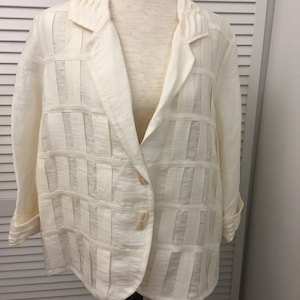 May include: A white linen jacket with a unique design featuring vertical and horizontal pleats. The jacket has a single button closure and long sleeves.
