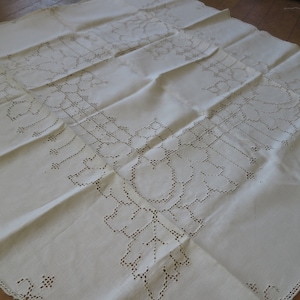 Puede incluir: Un mantel de lino blanco con un delicado diseño bordado. El bordado presenta un patrón floral con intrincados detalles.