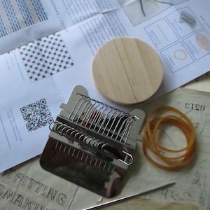 May include: A close-up of a loom kit for weaving. The kit includes a wooden loom, a metal weaving tool, a rubber band, a needle, and instructions. The instructions include a diagram and a QR code.