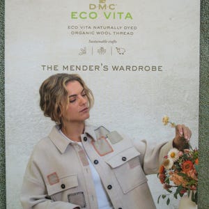 Può includere: Una copertina di libro con una donna che indossa una giacca beige con toppe colorate. Il titolo del libro è "The Mender's Wardrobe" e il sottotitolo è "21 Upcycling projects/21 proyectos de upcycling/21 progetti di upcycling". Il libro tratta di artigianato sostenibile utilizzando filo di lana biologico tinto naturalmente DMC Eco Vita.