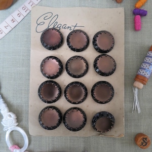 May include: A set of eleven round buttons arranged on a rectangular card labeled Elegant. Each button features a smooth, metallic bronze-toned center surrounded by a dark, scalloped rim. The card is positioned near a measuring tape showing inches.