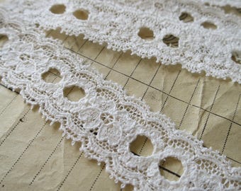 May include: Three pieces of white cotton lace trim with a delicate floral pattern and scalloped edges. The lace has small, round holes throughout the design.