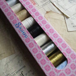 Può includere: Una scatola rosa con un motivo bianco e rosa. La scatola contiene otto bobine di filo di vari colori, tra cui bianco, beige, nero, argento e oro.