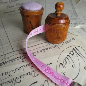 May include: Two wooden pincushions with a pink fabric tape measure. The pincushions are both round and have a knob on top. The tape measure is wrapped around the pincushions and has the word "SAJOU" printed on it.