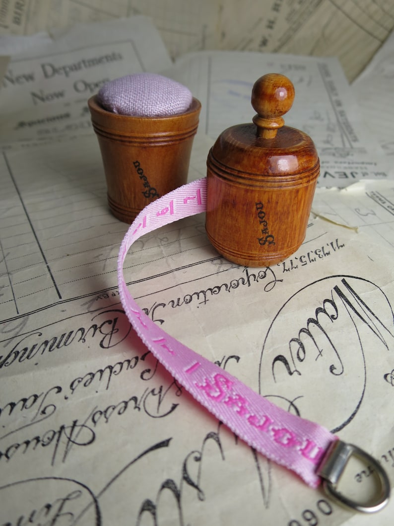 May include: Two wooden pincushions with a pink fabric tape measure. The pincushions are both round and have a knob on top. The tape measure is wrapped around the pincushions and has the word "SAJOU" printed on it.