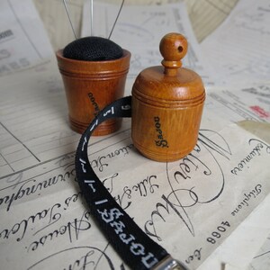 May include: Two wooden sewing accessories, a pincushion and a tape measure, with a black fabric top and a black and white tape measure. The pincushion has three silver pins sticking out of it. The tape measure has the word "SAJOU" printed on it.