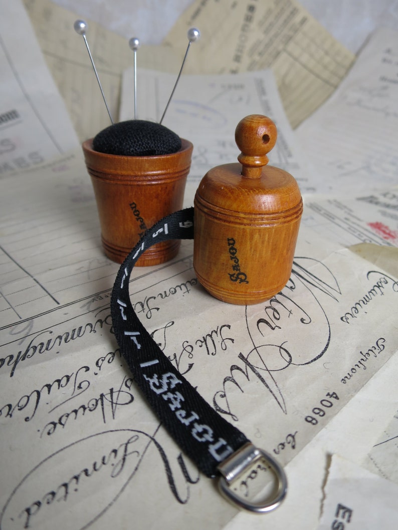 May include: Two wooden sewing accessories, a pincushion and a tape measure, with a black fabric top and a black and white tape measure. The pincushion has three silver pins sticking out of it. The tape measure has the word "SAJOU" printed on it.