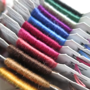 Può includere: Primo piano di una collezione di rocchetti di filo da ricamo in vari colori, tra cui giallo, marrone, rosso, blu, viola, verde e nero. I rocchetti sono disposti in fila, con i colori che sfumano dal chiaro allo scuro.