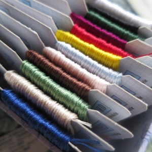 May include: A close-up of a variety of embroidery floss spools organized in a box. The floss is arranged in rows of different colors, including blue, green, white, brown, pink, yellow, red, purple, and black.
