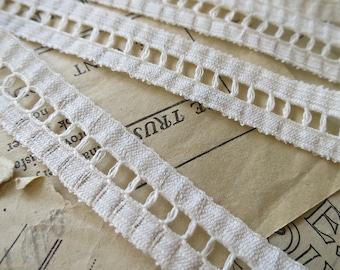 Puede incluir: Borde de encaje de algodón blanco con un delicado diseño calado. El encaje está dispuesto en varias capas sobre un fondo de papel texturizado.